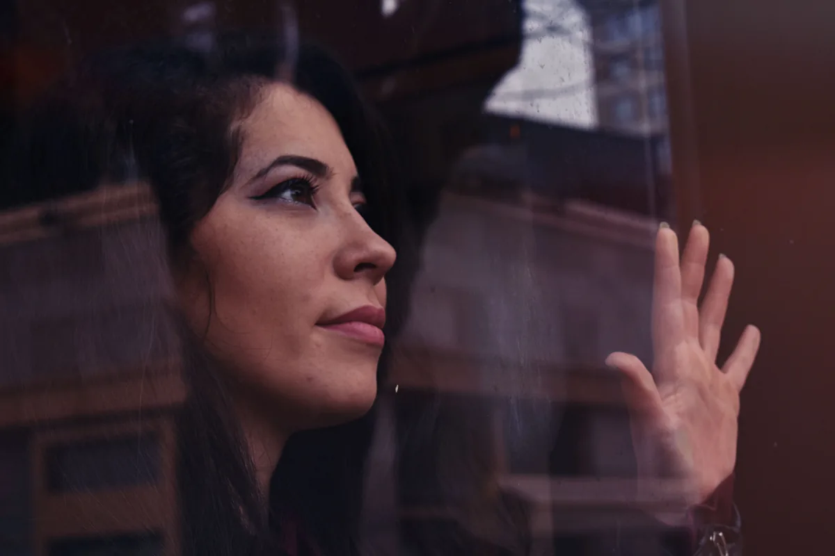 Woman looking out of a window on a grey autumn day, her hand resting on the glass: as the days get shorter, seasonal depression can bring your mood down