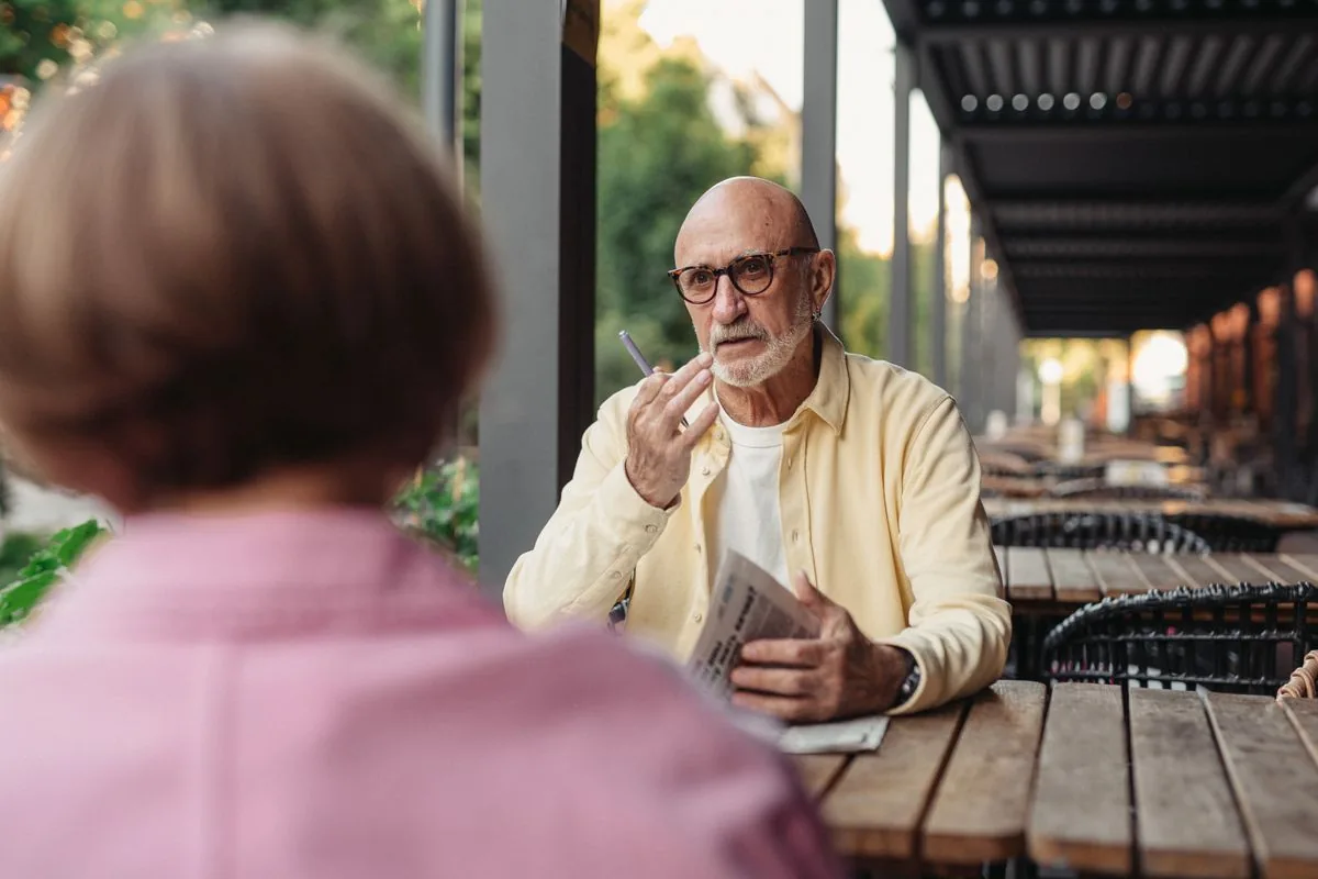 Older man with glasses on a café terrace listening closely to his companion, a noisy setting where hearing loss shows up first